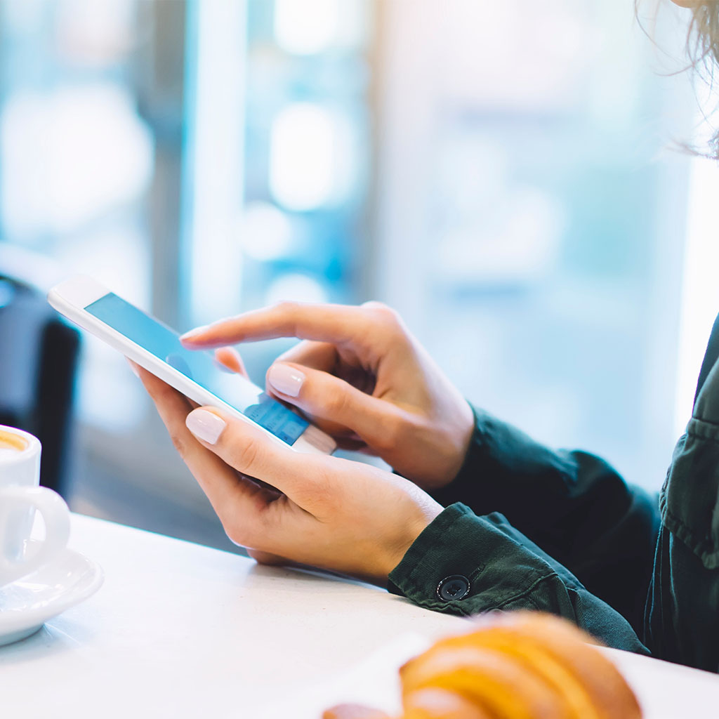 Woman tapping a smartphone at a cafe table, representing a customer browsing and converting on mobile.